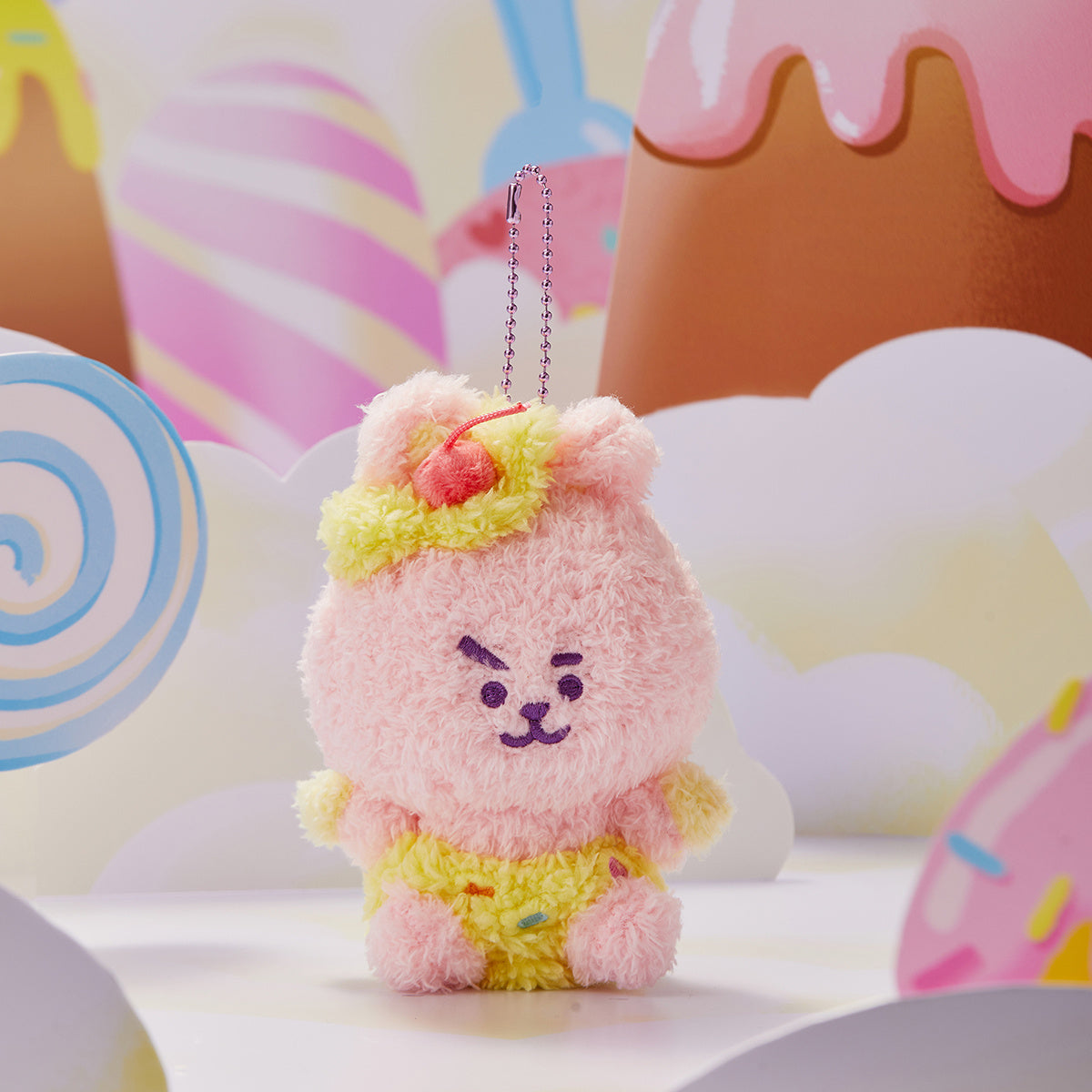 Pink plush bear keychain with yellow diaper and cherry on head, set in a candy-themed scene.