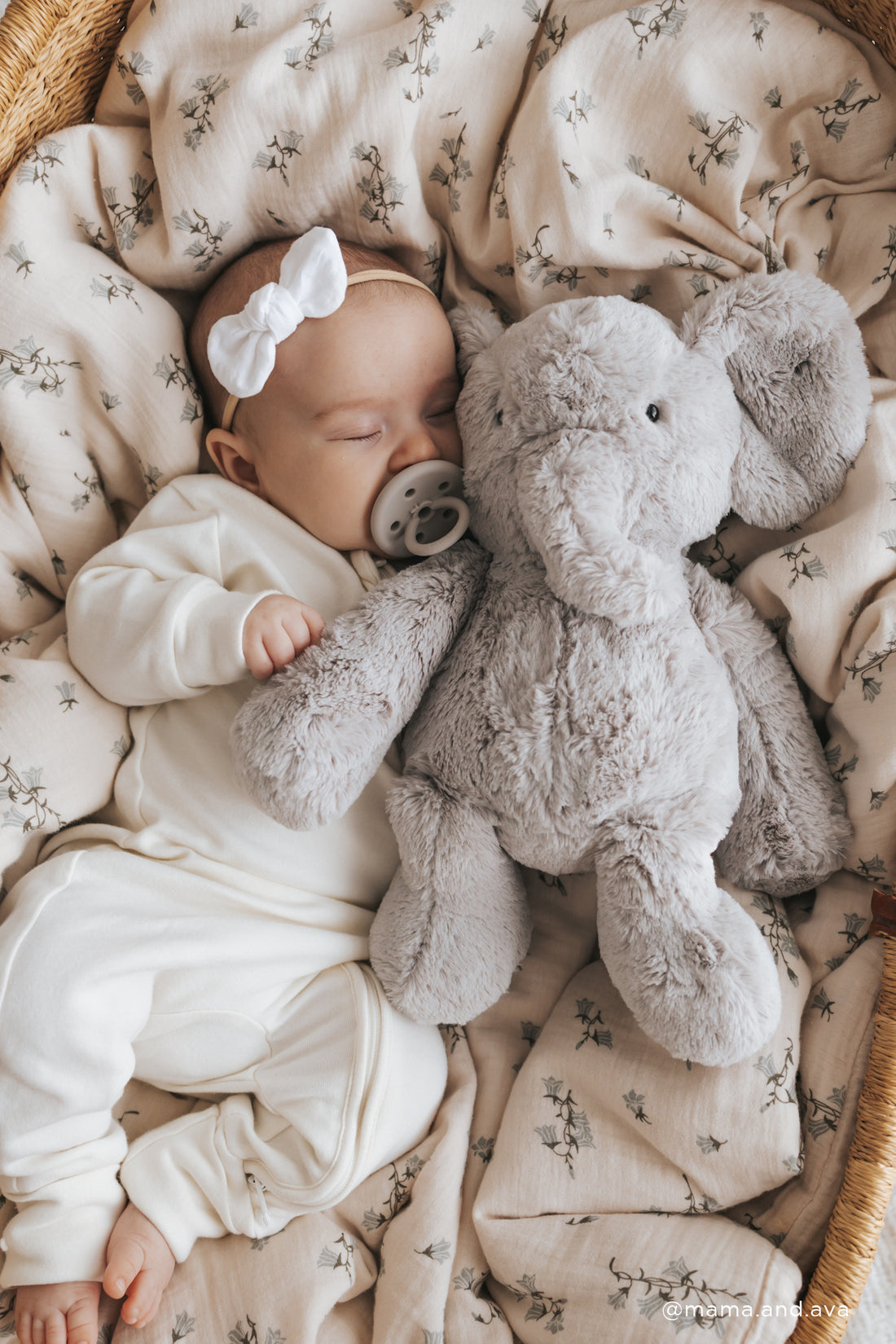 Sleeping baby with a white bow, pacifier, and plush gray elephant on a floral patterned blanket in a wicker basket.