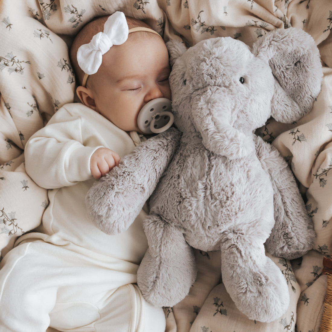 Sleeping baby with a white bow, pacifier, and plush gray elephant on a floral patterned blanket in a wicker basket.