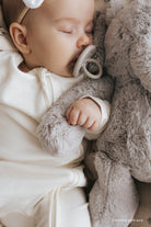 Close-up of a sleeping baby with a pacifier, wearing a white onesie, and cuddling a gray plush toy.