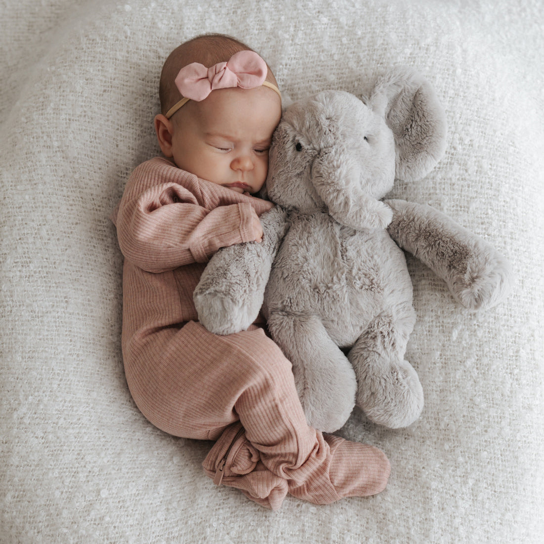 Sleeping baby with pink bow and onesie, cuddling a gray stuffed elephant on a white blanket.