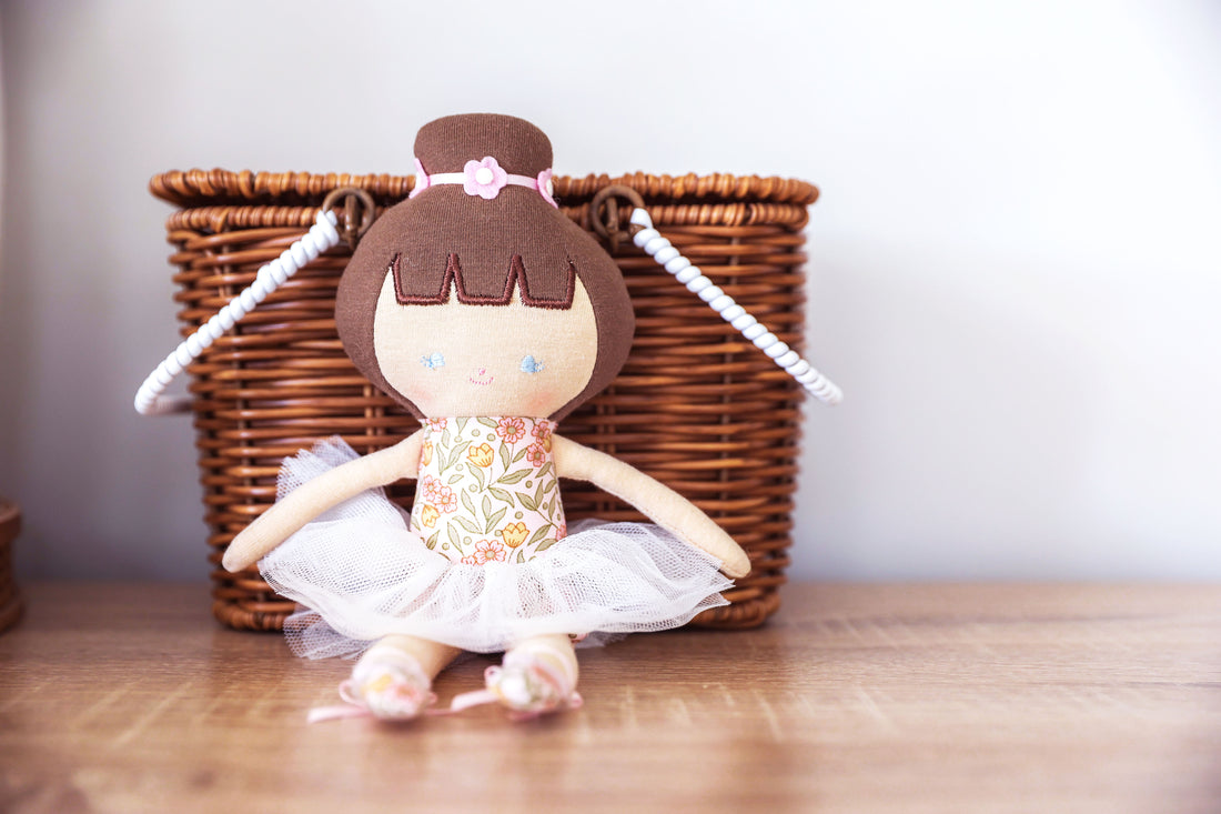 Stuffed ballerina doll with brown hair, floral dress, and white tutu sitting on a wooden surface in front of a wicker basket.