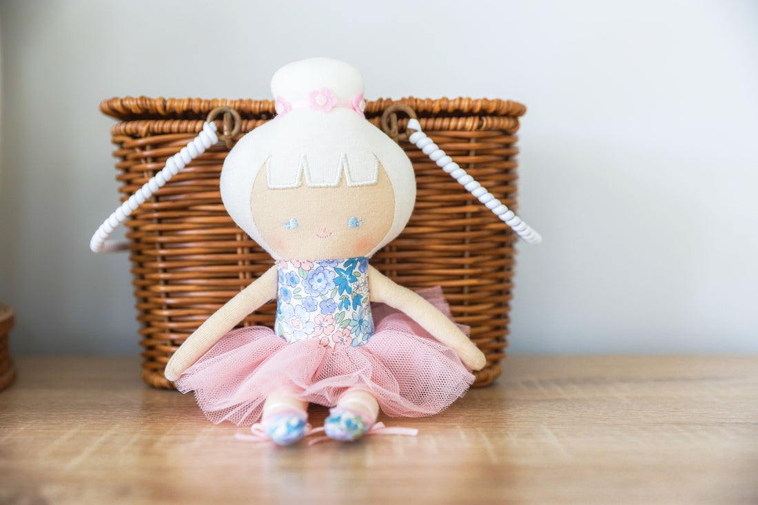 Ballerina doll in a pink tutu, floral dress, and ballet shoes, sitting in front of a wicker basket.