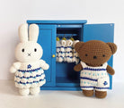 Two crocheted stuffed toys, a bunny and a bear, stand on either side of a blue cabinet. The bunny wears a white dress with blue trim, and the bear wears white overalls with blue stripes. Clothes hang inside the cabinet.