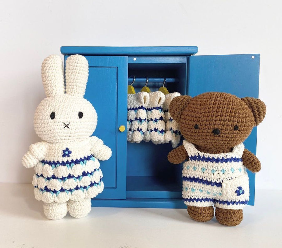 Two crocheted stuffed toys, a bunny and a bear, stand on either side of a blue cabinet. The bunny wears a white dress with blue trim, and the bear wears white overalls with blue stripes. Clothes hang inside the cabinet.