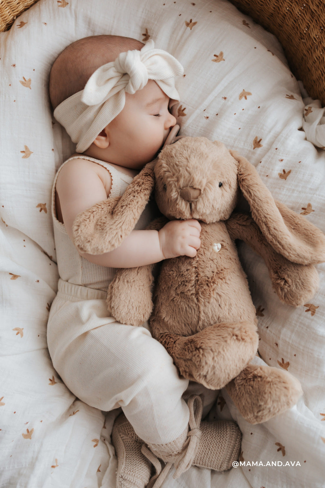 Sleeping baby with headband hugging a brown plush bunny in a patterned baby bed.