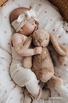 Sleeping baby with headband hugging a brown plush bunny in a patterned baby bed.