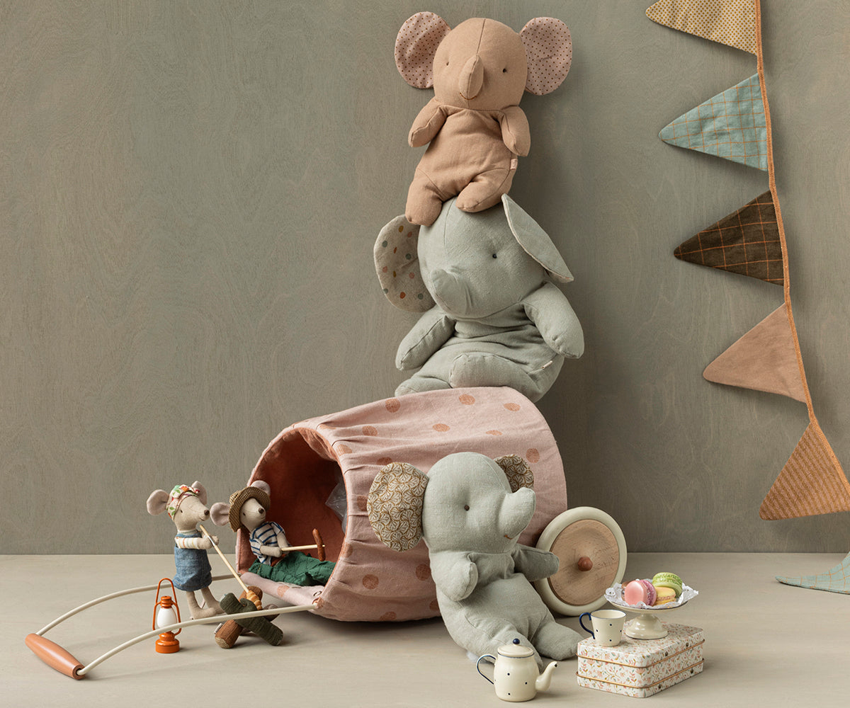 Stuffed elephants stacked next to a polka-dot toy tunnel with toy mice, a toy campfire, and a toy tea set with macarons. There is also a banner of triangular flags.