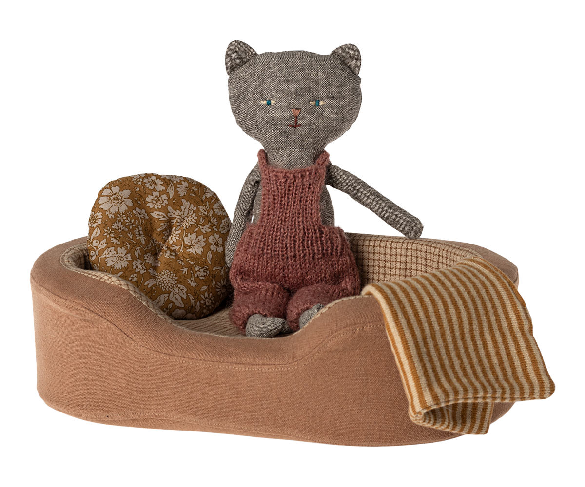 Gray cat doll in red overalls in brown bed with pillow and striped blanket.