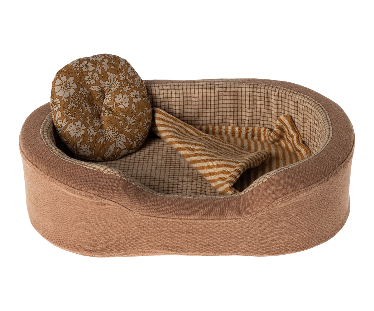 Oval, brown dog bed with floral pillow and striped blanket.