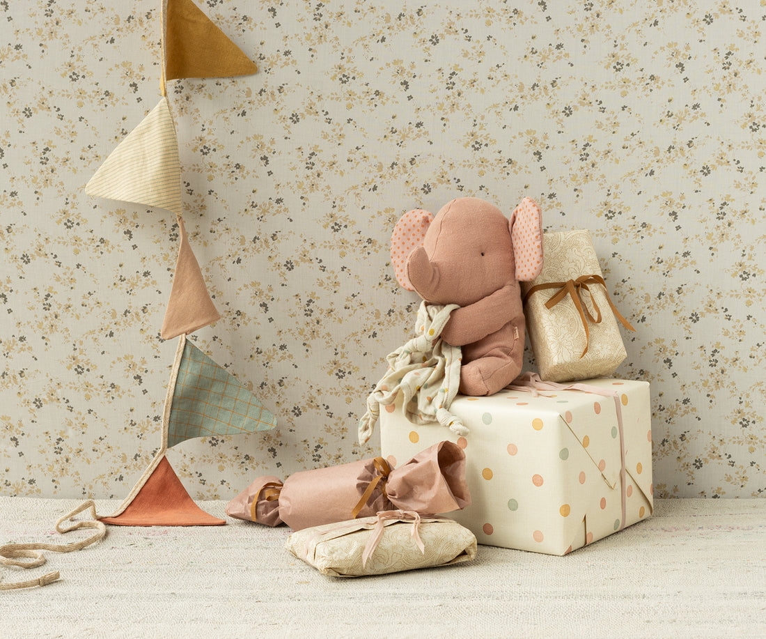 Close-up of children's toys and gifts against a floral wallpaper. A plush pink elephant sits atop a polka-dotted gift box next to a gift wrapped in beige paper, other small gift wraps around on the beige carpet. A colorful triangular banner hangs on the wall.