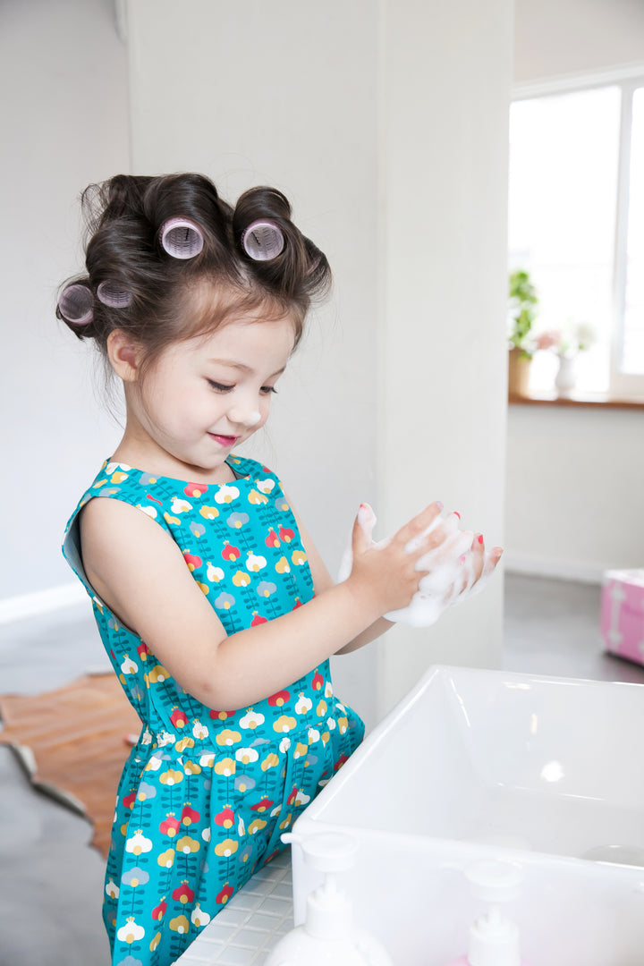 Child in hair rollers and patterned dress washing hands with soap.