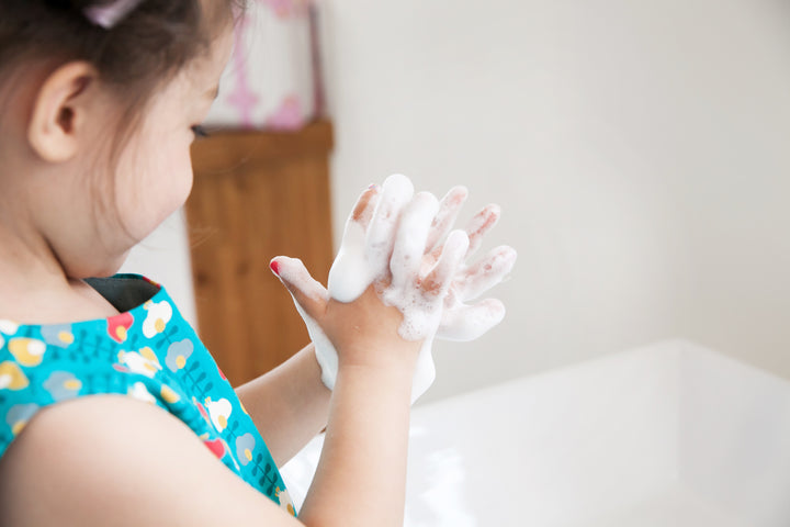 Young child washing hands with soap. Child wears a blue and green floral dress.