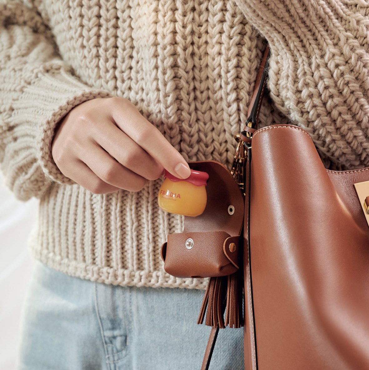 Woman holding Frudia honey lip balm attached to a brown leather purse while wearing a beige knitted sweater.