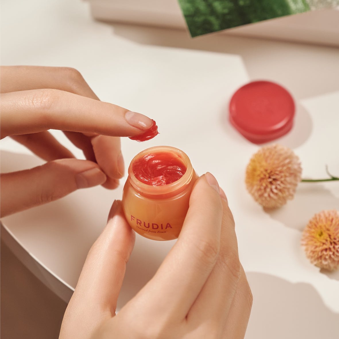 Hands holding a small orange jar of Frudia lip balm with red lip balm on a finger, red lid and flowers on a cream colored surface.