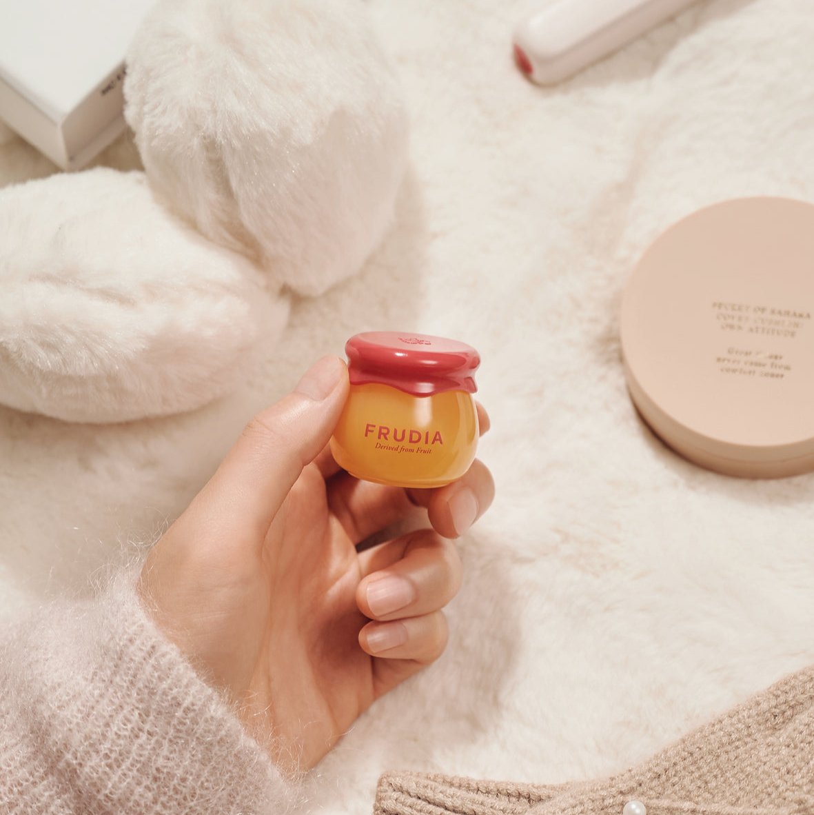 A hand holding a small, jar-shaped container of Frudia lip balm against a soft, textured background.