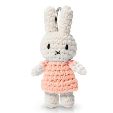 Here are a few alt tag options for the image, focusing on different details:
*   **Crocheted Miffy rabbit doll with peach dress and key ring.**
*   **Small cream-colored yarn Miffy keychain toy.**
*   **Front view of crochet Miffy wearing a pink dress.**