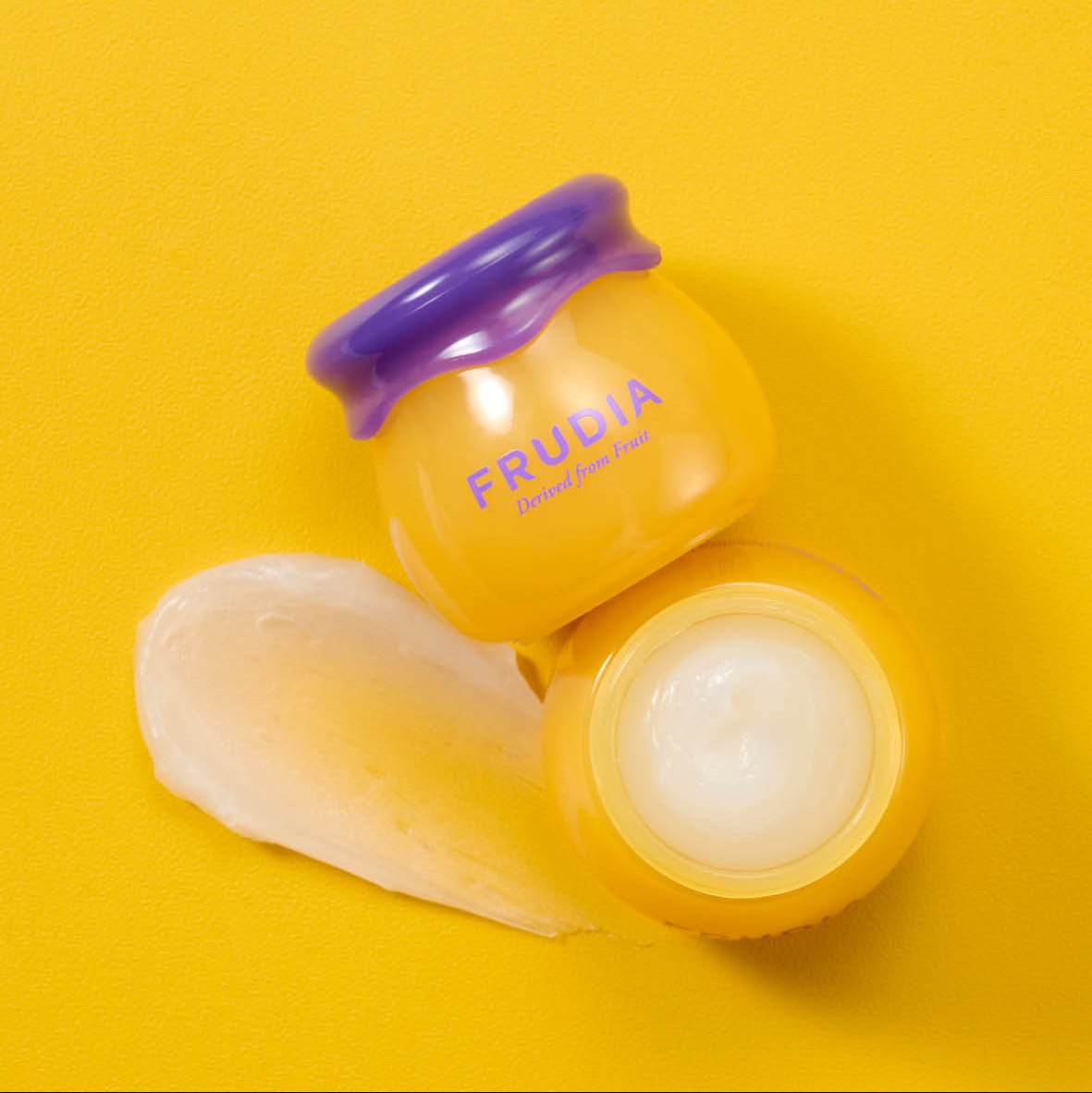 Two yellow Frudia jars, one open, with a white smear of cream on a yellow background.