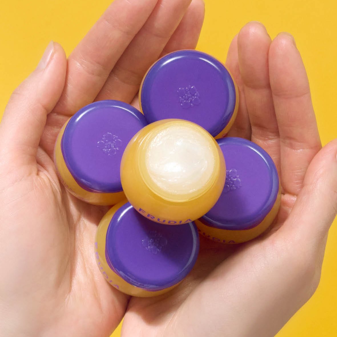 Hands holding five Frudia lip balms with yellow bases and purple lids.