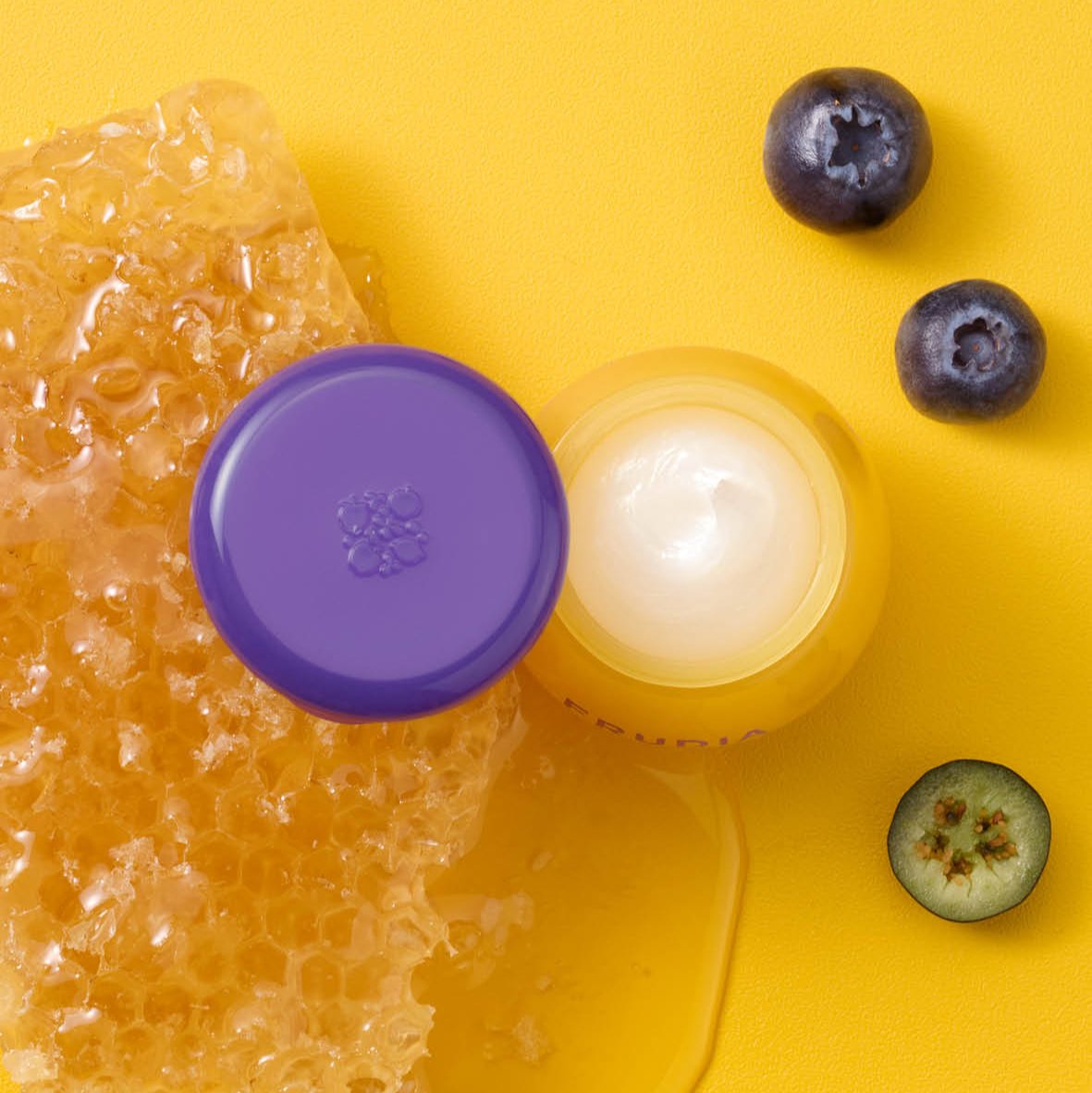 Overhead shot of skincare product, honey, and blueberries against yellow background.