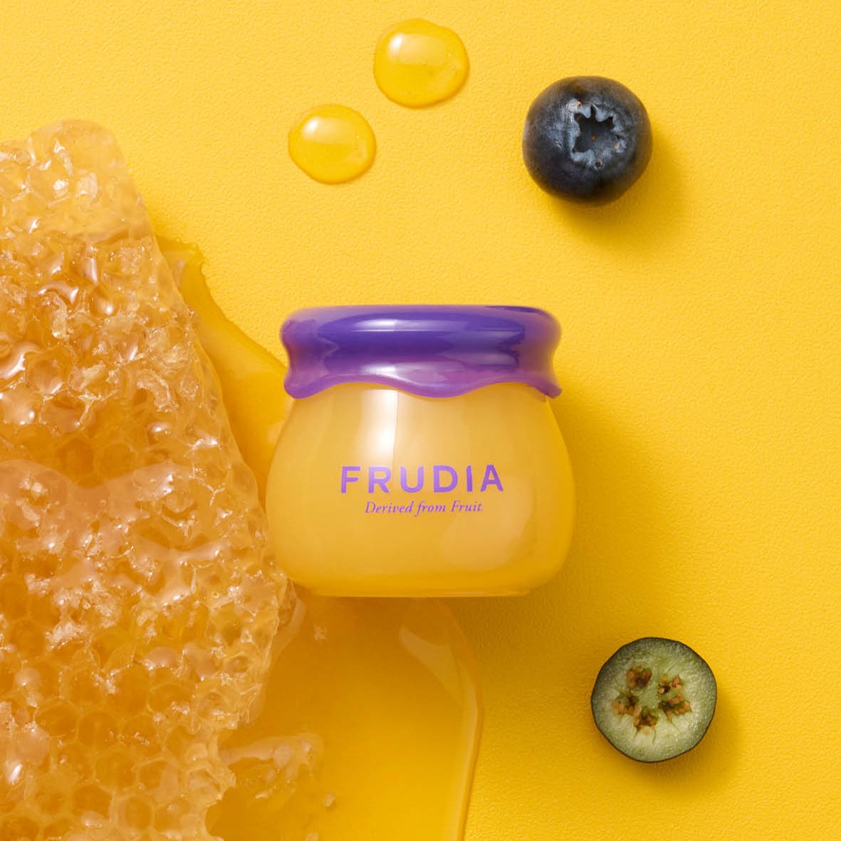 Overhead shot of a jar of Frudia skincare product next to honeycombs, drops of honey, and blueberries, all on a yellow background.