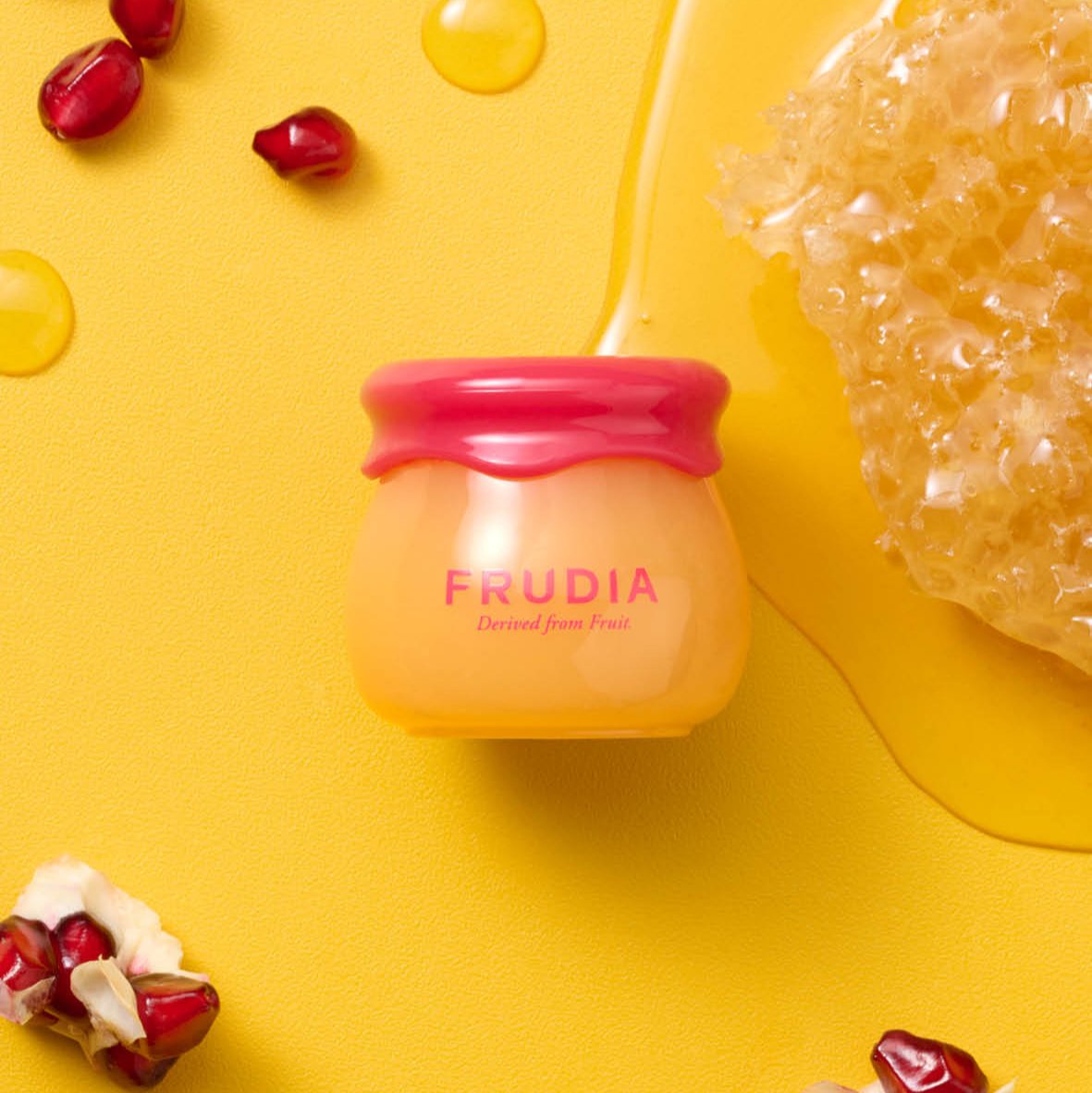 Close-up of a Frudia jar on a yellow surface, with pomegranate seeds and honeycomb.
