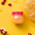 Close-up of a Frudia jar on a yellow surface, with pomegranate seeds and honeycomb.