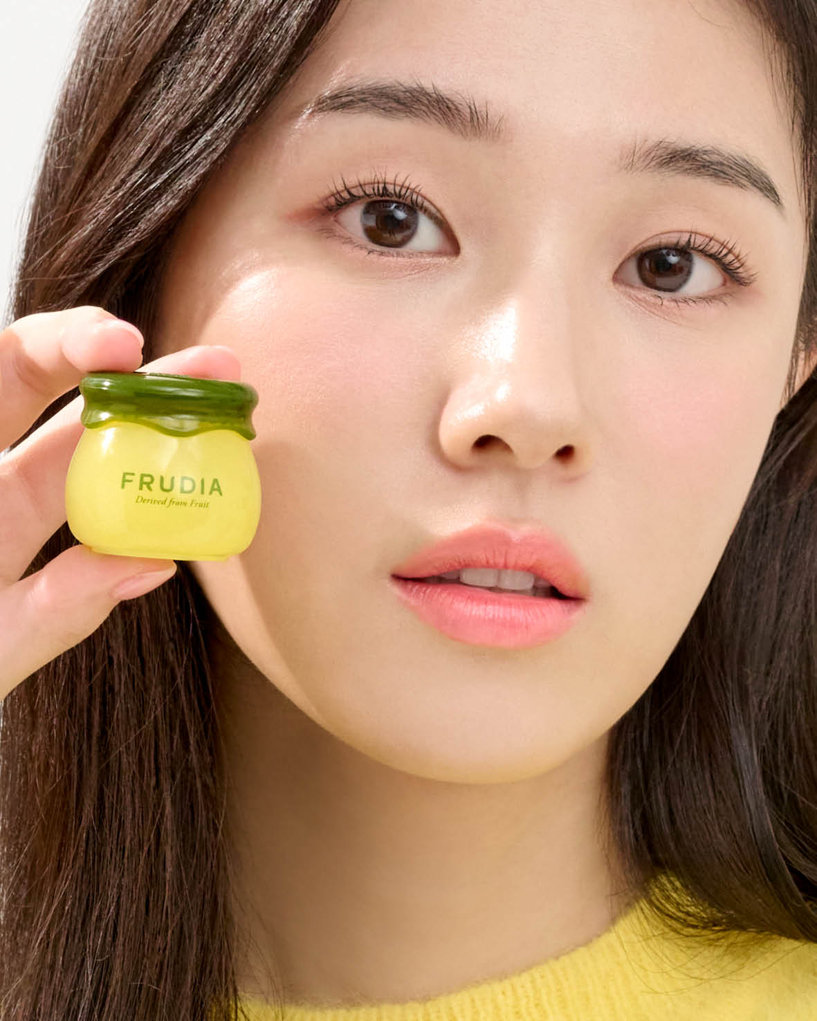 A woman holds a small yellow jar of Frudia skincare product.