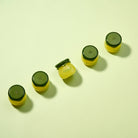 Five small yellow Frudia jars with dark green lids arranged diagonally on a light green background.