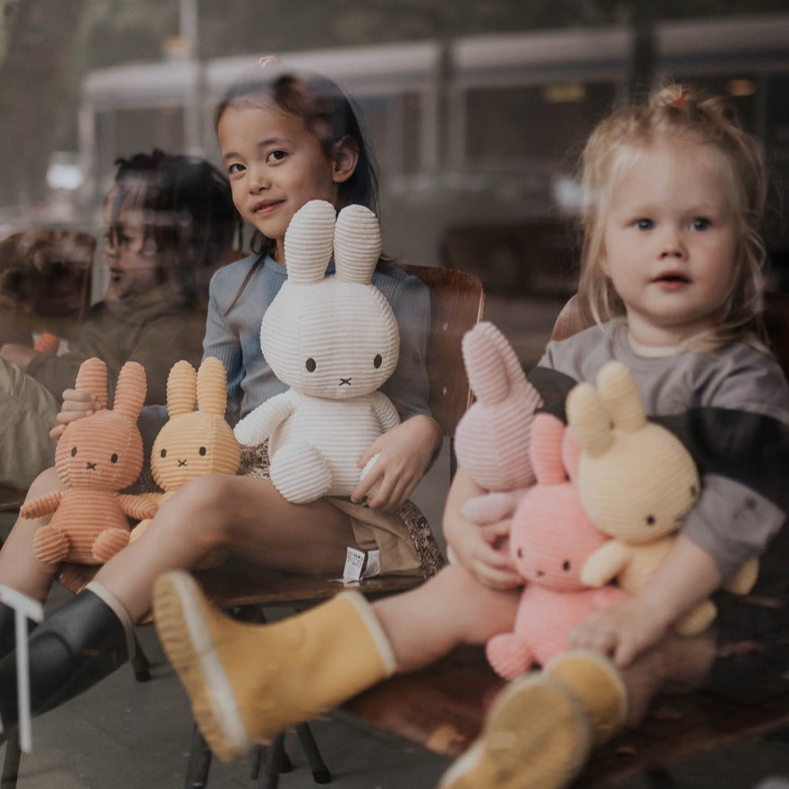 Two young children sitting on chairs holding plush Miffy bunnies of different colors.