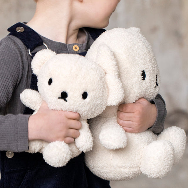 Child holding a white stuffed bear and a white stuffed bunny. The child is wearing a gray long-sleeved shirt and dark blue overalls.
