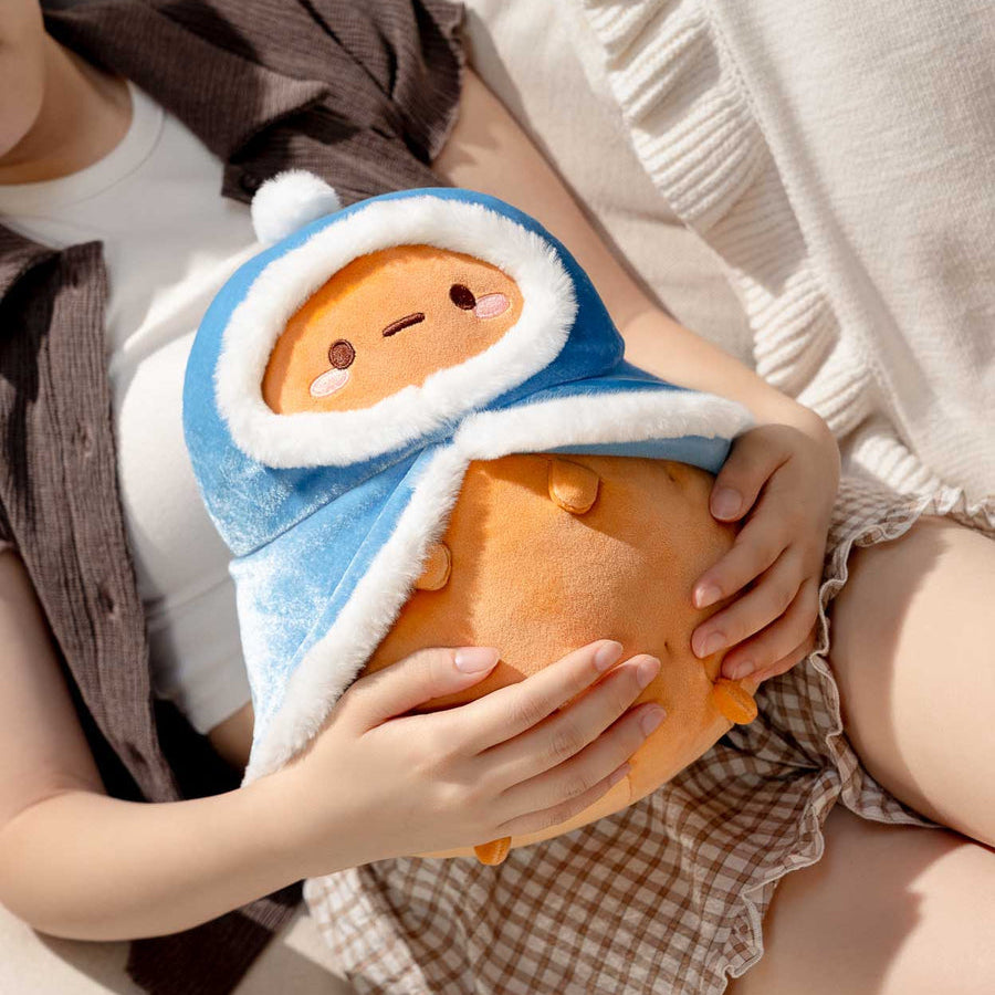 A person reclining on a beige sofa and holding a plush toy wrapped in a blue hood with white faux-fur trim. The toy has an orange body and a cute face.
