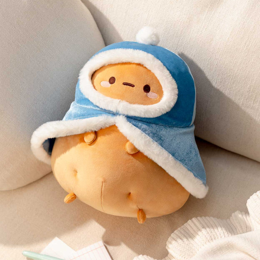 A cute potato-shaped plush toy wearing a blue winter hat with white trim, styled on a beige sofa with pillows and a knit blanket nearby.