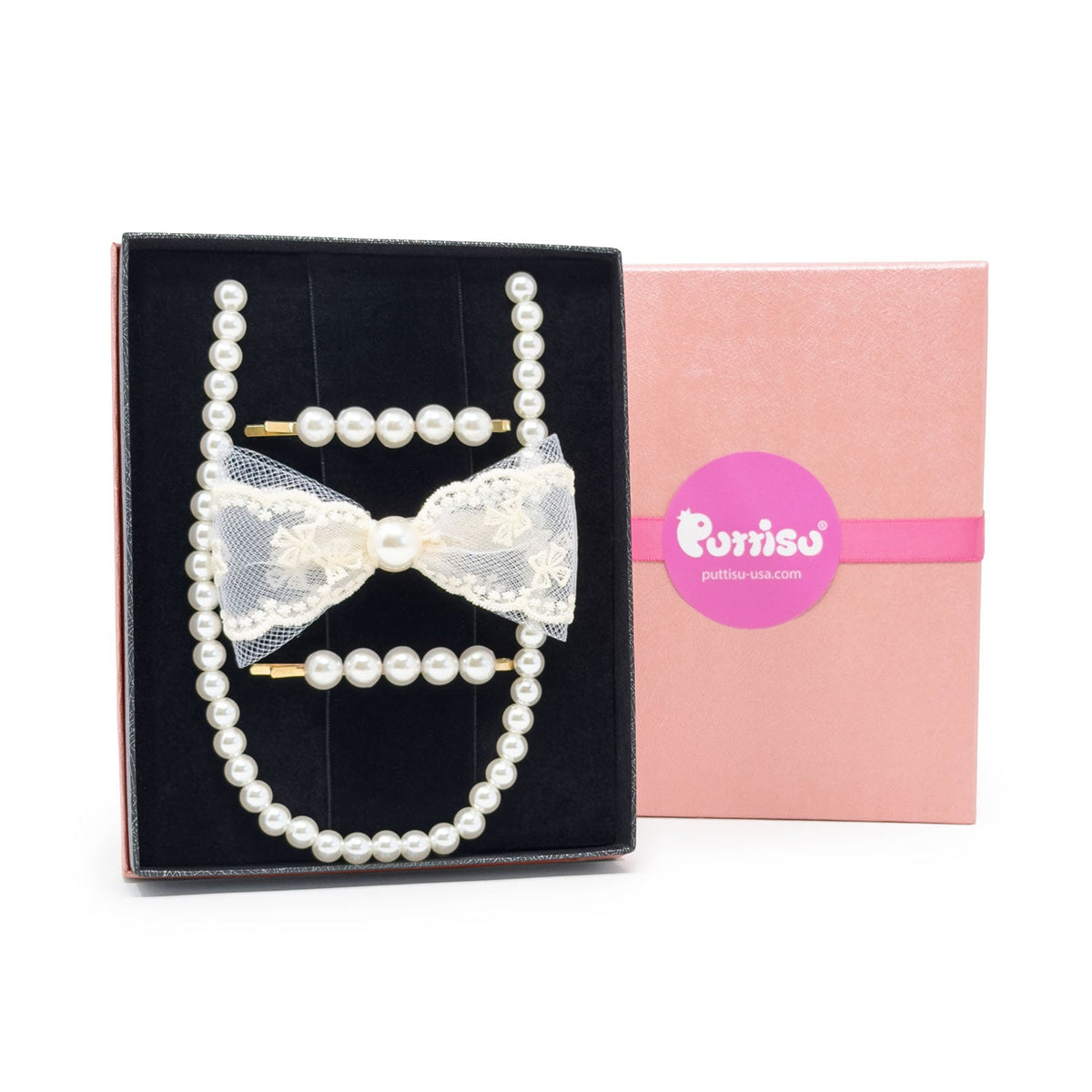 A pearl hair accessory set displayed in a black presentation box next to its pink branded box. The set includes a long pearl strand, two pearl hair clips with gold-tone ends, and a white lace bow with a central pearl.