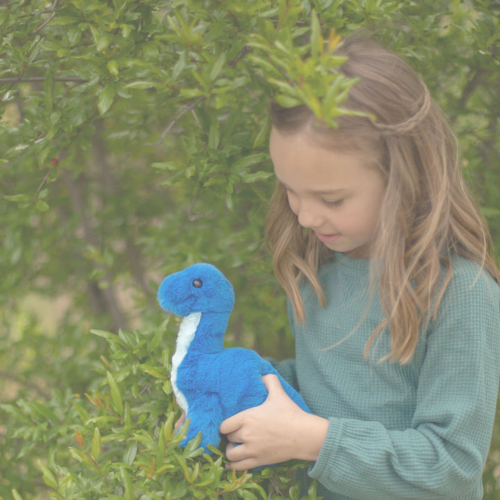 Young girl holding a blue, plush dinosaur toy in a leafy outdoor setting.