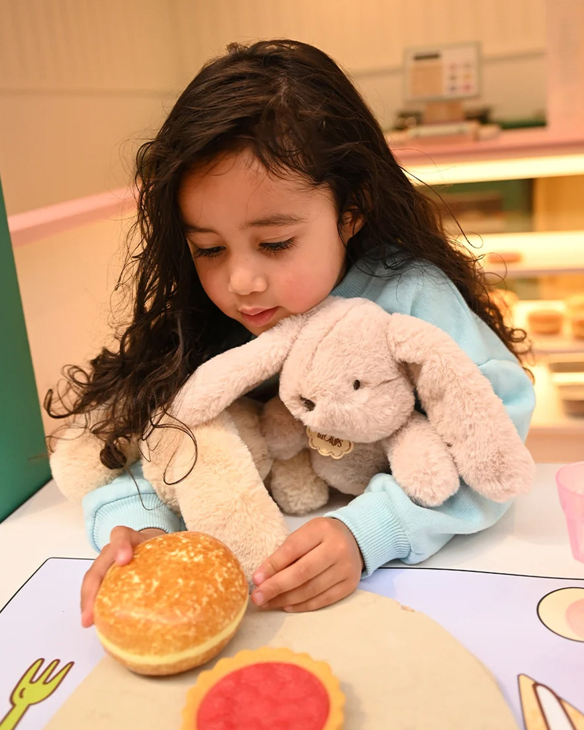 Child holding a toy hamburger with a stuffed animal, sitting at a table with a colorful mat.