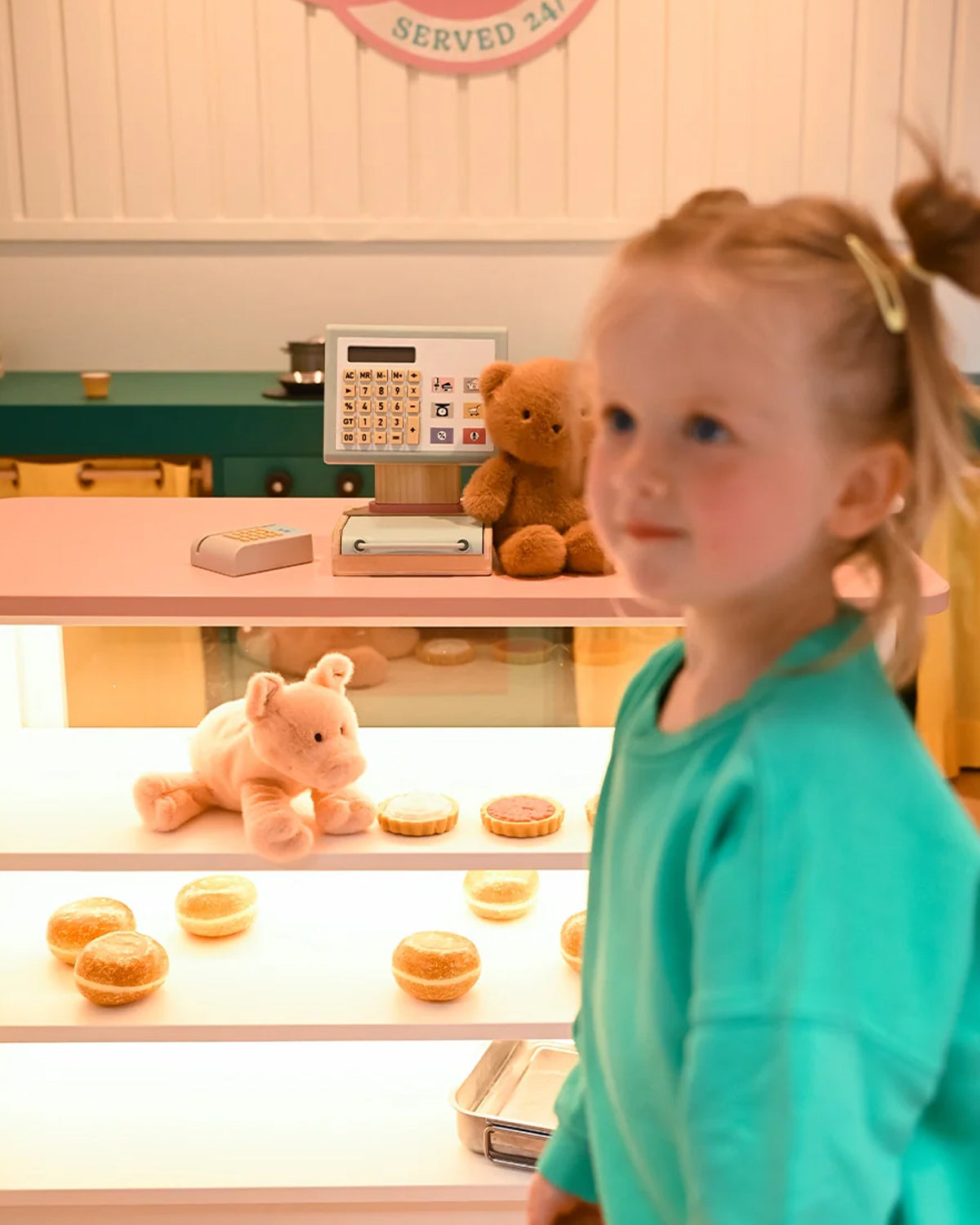 Young girl in a teal shirt standing in front of a toy cash register with teddy bears and donut toys on shelves.