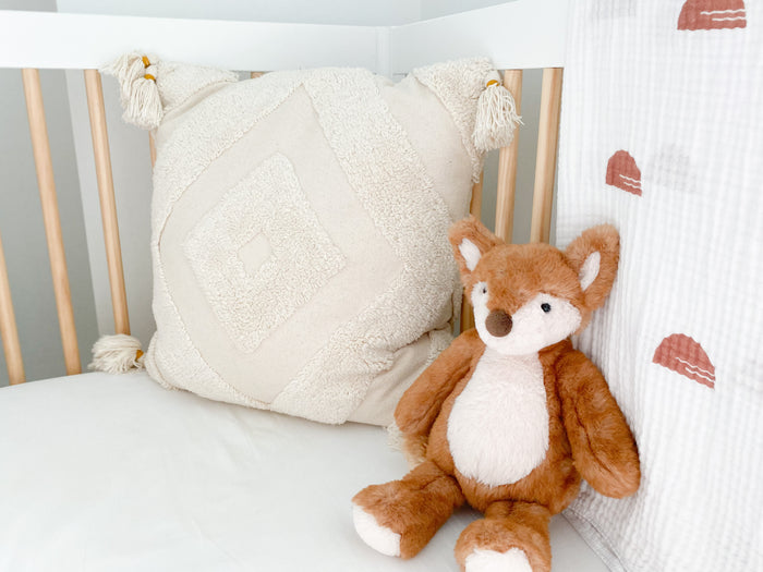 Tan tasseled decorative pillow and fox plush in a crib.