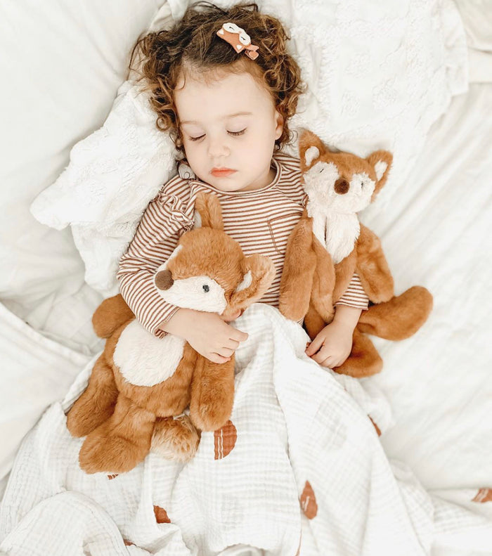 Toddler sleeping with stuffed foxes in a brown and white striped top, white blanket and fox hair clip.