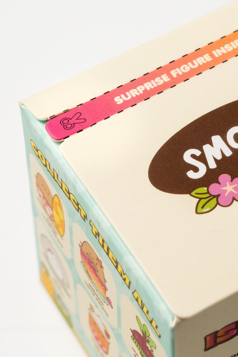 Close-up of a collectible toy box showing a pink flap sticker that reads 'SURPRISE FIGURE INSIDE' and part of the brand name 'SMO' on a cream-colored box with pastel illustrations.