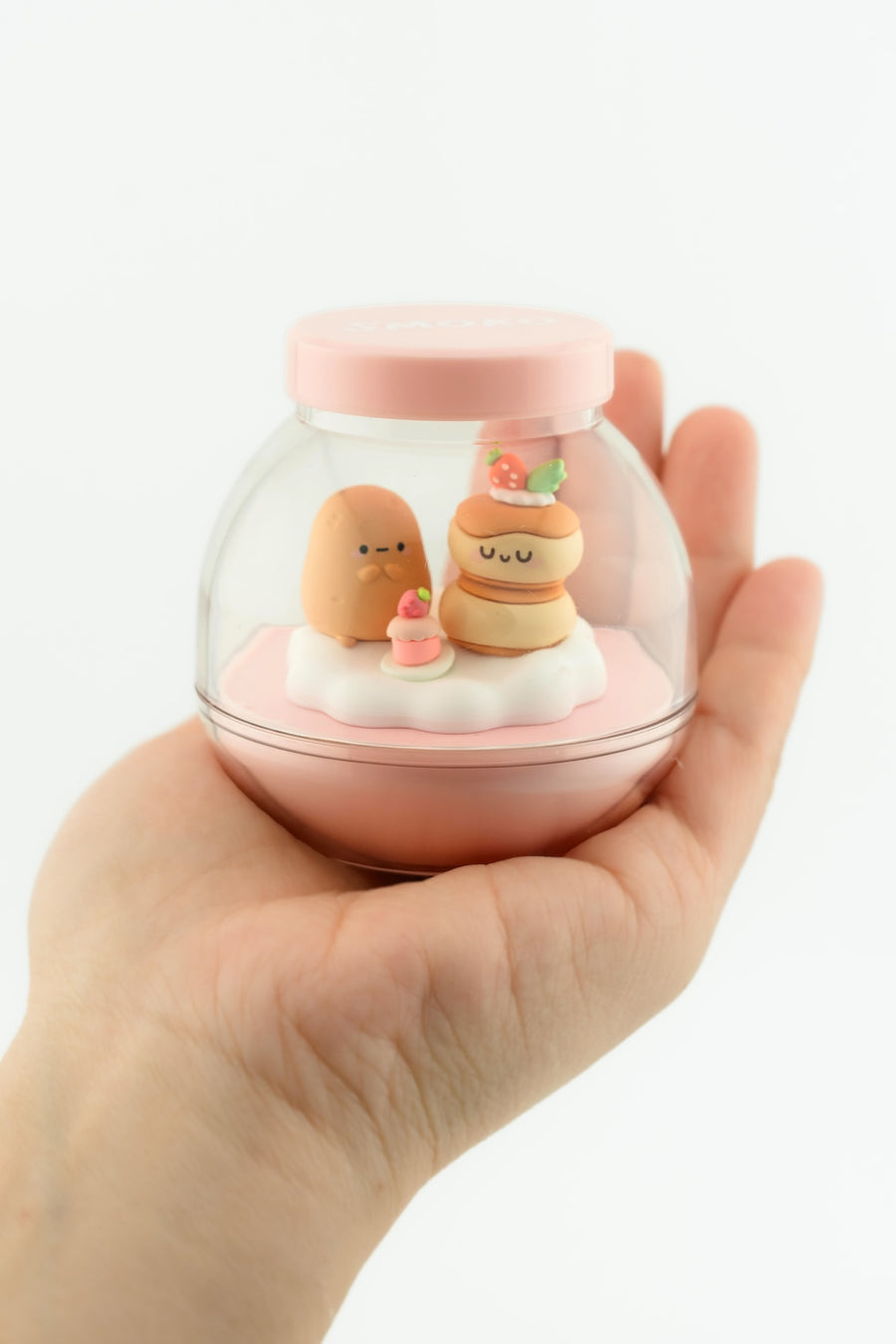 Hand holding a small glass container with decorative food-themed figures on a white background