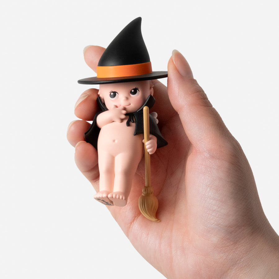Halloween-themed baby figurine with a witch hat and broomstick held in a hand against a white background