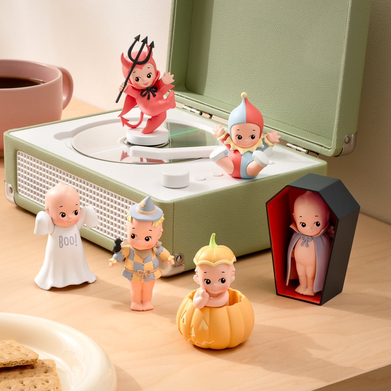 A styled tabletop scene with a sage green record player as the main product, surrounded by small decorative figurines and props including a cup, plate, and a coffin-shaped display box.