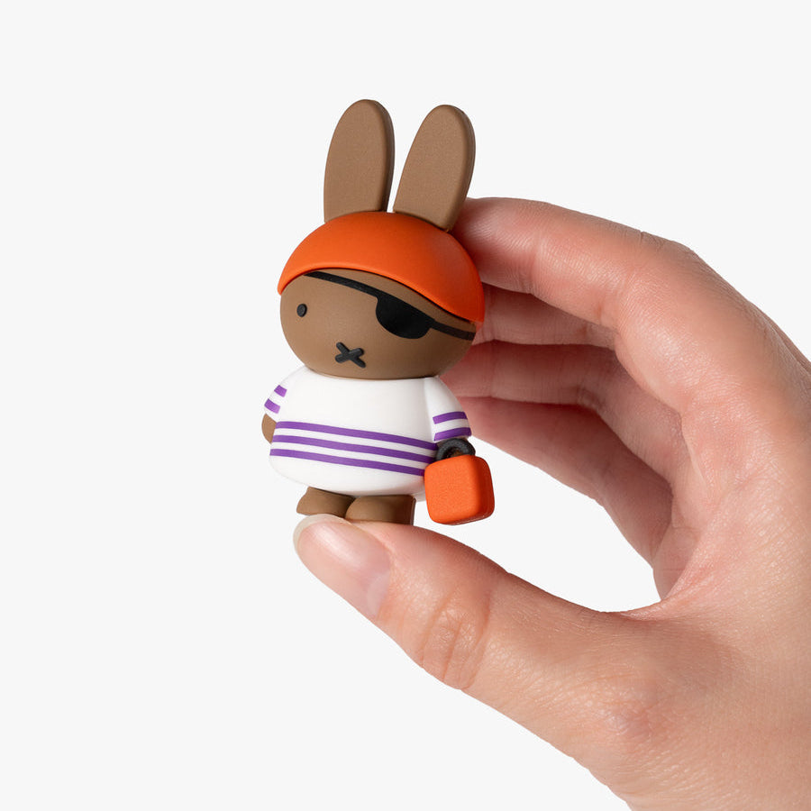 A small cartoon rabbit figurine with long ears, wearing an orange pirate hat, black eye patch, white shirt with purple stripes, and holding an orange bag, being held by a human hand against a plain white background.