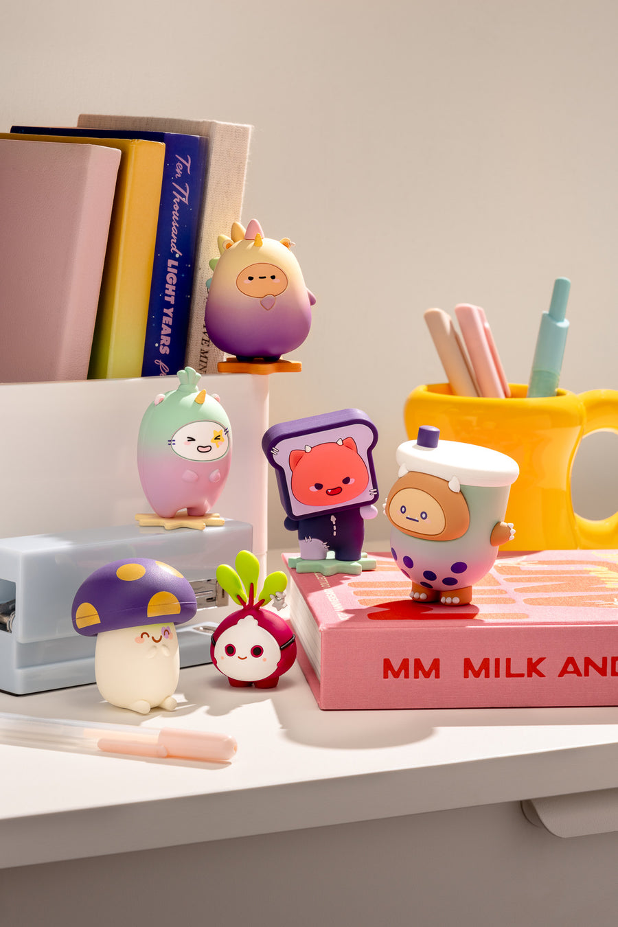 A collection of cute, colorful cartoon desk accessories arranged on a white desk, including a mushroom-shaped pencil holder, a radish-shaped eraser, a cup-shaped highlighter, and egg-shaped desk lights, alongside books and a yellow pen cup in the background.