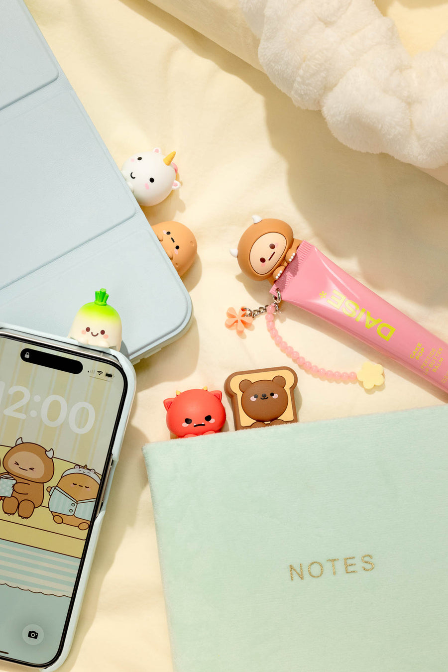 A styled flat-lay of cute cartoon accessories on a cream fabric background, including a light blue phone case, a mint green notebook with gold 'NOTES' text, a pink pen, and several small bear-shaped charms.