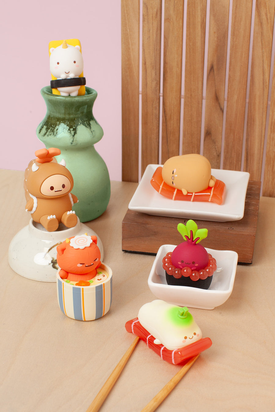 A set of cute, food-themed silicone bath toys shaped like sushi, a dumpling, and a character on a chopstick, displayed alongside a green ceramic vase and wooden blocks on a light wood surface.