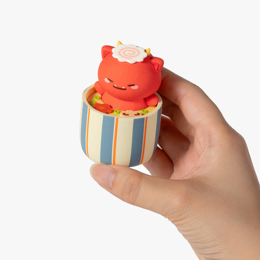 A hand holding a small decorative figurine shaped like a red cat sitting inside a striped cup filled with yellow items, against a plain white background.