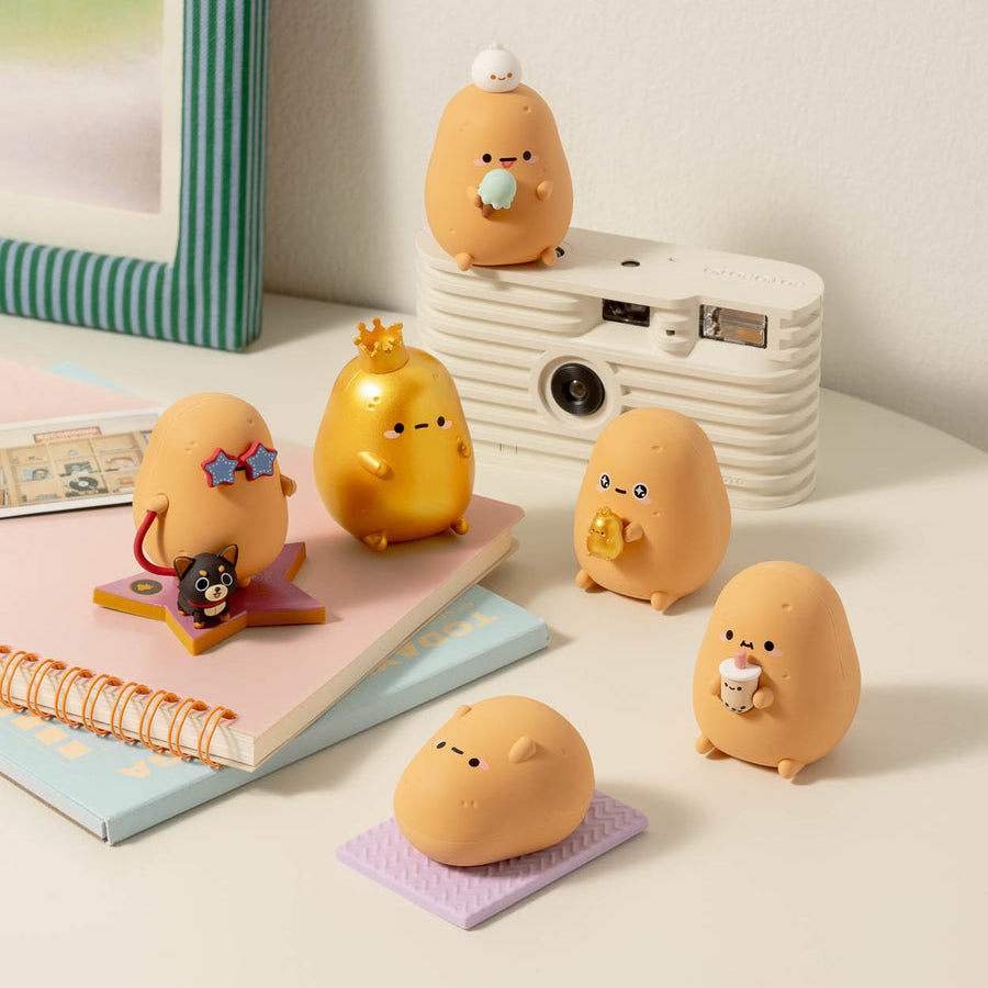 A set of cute, egg-shaped character figurines in various poses and colors, styled on a white table next to a retro camera, notebook, and striped frame.