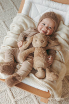 Baby in brown outfit and hat, holding a stuffed bunny, laying on a white blanket in a wooden cradle.